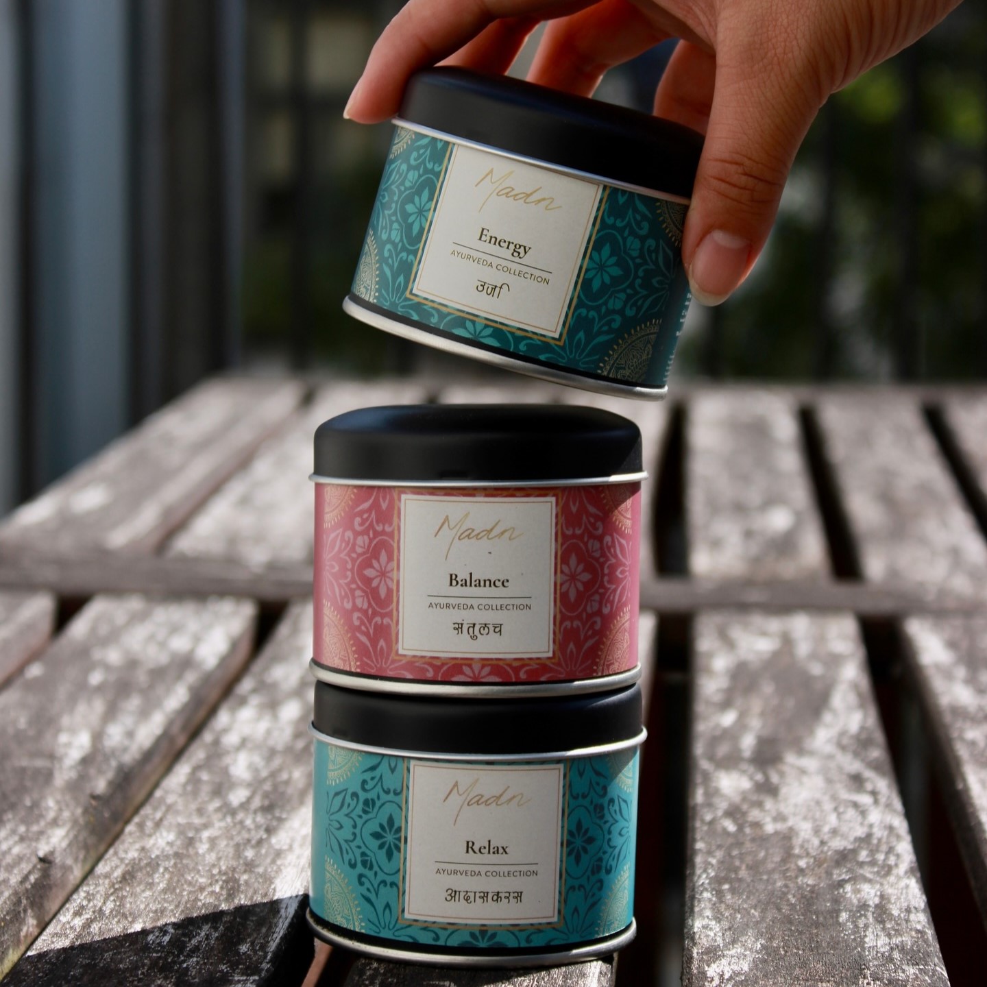 Three Madn Ayurveda Collection tea tins — Energy, Balance, and Relax — stacked on a weathered wooden table, with a hand placing the Energy tin on top in natural outdoor light.